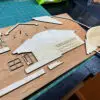 Workbench scene with plywood cutouts and thin wooden strips scattered on a wooden surface, a green cutting mat and measuring tape in the background